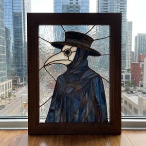 Könnte beinhalten: Ein Buntglasfenster in einem dunklen Holzrahmen zeigt einen Pestdoktor mit breitkrempigem Hut und langschnäbeliger Maske. Die Figur ist aus blauem und weißem Glas mit dunklen Lötlinien gefertigt und vor einem strukturierten, klaren Hintergrund.
