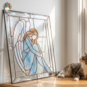 May include: Stained glass panel depicting an angel in a seated position, wearing a light blue dress and iridescent wings. The artwork is framed in a rectangular shape with a clear glass border. A cat is lying down next to the panel.