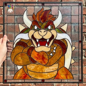 May include: A square stained glass panel featuring a depiction of the character Bowser with orange, brown, and cream glass pieces held together by black lead cames. The character has horns, a spiked shell, and a fierce expression. Text reads Punshop.