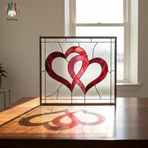 Op de afbeelding: Een glas-in-lood paneel met twee verweven rode harten. Het paneel is omlijst in donker metaal en staat op een houten oppervlak. Het zonlicht werpt een hartvormige schaduw. Afmetingen ca. 25 cm x 25 cm.