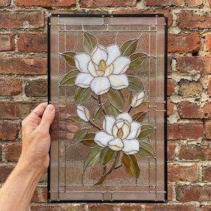 Puede incluir: Un panel rectangular de vidrieras que presenta dos flores de magnolia blancas con centros amarillos y hojas verdes. El diseño está enmarcado por un borde de metal oscuro con paneles de vidrio transparente texturizado en los bordes.