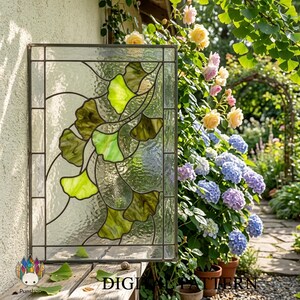 May include: Rectangular stained glass panel featuring a ginkgo leaf design in shades of green and brown. The glass has a textured appearance and is framed with dark metal. The panel is displayed outdoors, with a garden in the background.