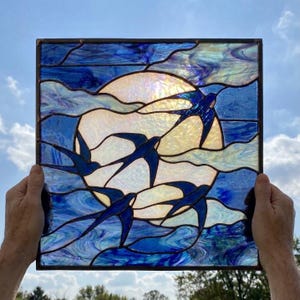 Puede incluir: Un panel cuadrado de vidrieras que presenta una gran luna circular blanca e iridiscente sobre un cielo azul profundo y arremolinado. Cuatro golondrinas azul oscuro vuelan frente a la luna y las nubes, unidas por líneas de soldadura metálica.