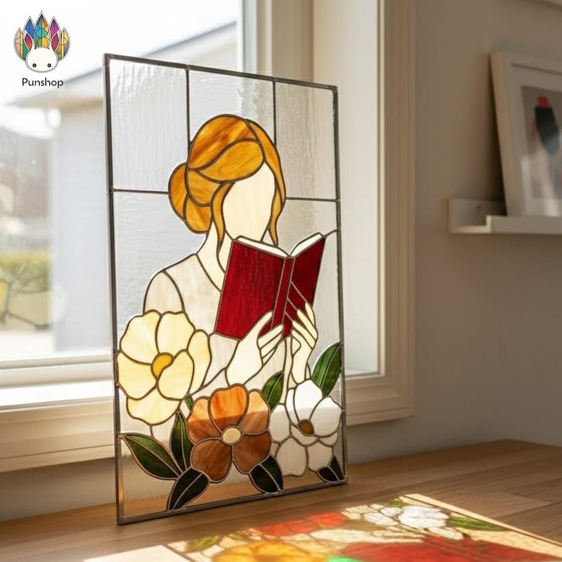 May include: Stained glass panel featuring a woman reading a book with a red cover. The woman has blonde hair and is surrounded by colorful flowers in shades of yellow, orange, and white. The panel is set against a window.