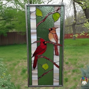 Peut inclure: Panneau en vitrail représentant deux cardinaux perchés sur une branche avec des feuilles vertes. Le panneau a un fond à rayures vertes et blanches. Le cardinal mâle est rouge vif, tandis que la femelle est brune. Le panneau est encadré en argent.