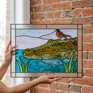 Op de afbeelding: Een rechthoekig glas-in-loodpaneel met een groene alligator die rust in blauw water met groen riet. Een kleine bruine vogel met een oranje borst zit op de kop van de alligator. De scène is geplaatst tegen een bleke lucht en omlijst door glas.