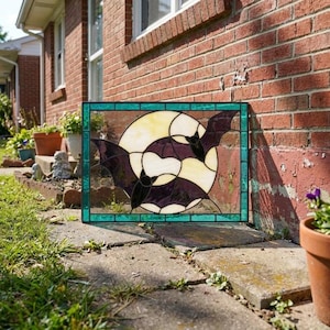 May include: A rectangular stained glass panel featuring two dark purple bats flying in front of a large, circular, pale yellow moon. The entire scene is framed by a border of small, rectangular, teal-colored glass pieces.