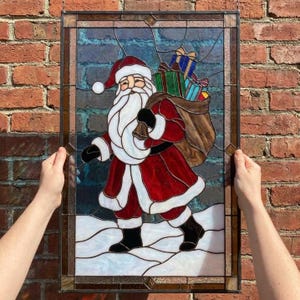 Peut inclure: Un panneau rectangulaire en vitrail représentant le Père Noël vêtu d'un costume rouge à bordure blanche, portant un sac marron rempli de cadeaux colorés. Le personnage se tient sur un sol enneigé blanc devant un fond bleu foncé.