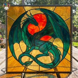 Op de afbeelding: Gebrandschilderd glaspaneel met een groene draak tegen een rode en oranje zon. De draak staat centraal in een cirkelvormig ontwerp, tegen een goudkleurige achtergrond en een vierkante rand.