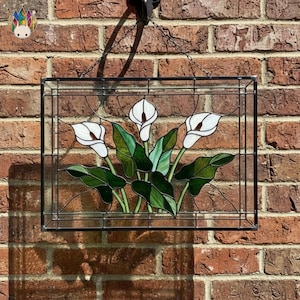 May include: Stained glass panel featuring white calla lilies with green leaves, set against a black frame. The artwork is suspended by a chain and hook. The panel is rectangular and hangs in front of a brick wall.