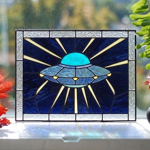May include: A rectangular stained glass panel featuring a central flying saucer in shades of blue and teal. The saucer emits yellow light rays against a dark blue background. The piece is framed by a clear, textured glass border.