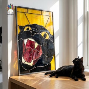 Könnte beinhalten: Ein Buntglasfenster mit einem schwarzen Panther mit offenem Maul, das rote und weiße Zähne und Zunge zeigt. Der Hintergrund ist gelb. Eine schwarze Katze liegt neben der Tafel auf einer Holzoberfläche.