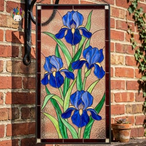 Peut inclure: Panneau en vitrail représentant des fleurs d'iris bleues avec des accents jaunes et des feuilles vertes. L'œuvre est encadrée d'une bordure marron foncé et suspendue sur un fond texturé, mettant en valeur un thème floral.