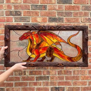 Könnte beinhalten: Ein rechteckiges Buntglasfenster mit einem Drachen in Rot-, Orange- und Gelbtönen. Der Drache hat einen langen, geschwungenen Schwanz und Flügel. Das Stück ist in einen dunkelbraunen, strukturierten Holzrahmen mit geschnitzten Augenmotiven gefasst.