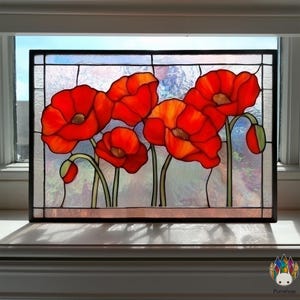 Op de afbeelding: Gebrandschilderd glaspaneel met levendige rode klaprozen met gele centra en groene stelen. De bloemen staan tegen een doorschijnende achtergrond, omlijst door een zwarte rand. Het paneel wordt getoond op een witte vensterbank.