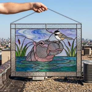 Peut inclure: Un panneau rectangulaire en vitrail représentant un hippopotame immergé dans une eau bleue avec une petite mésange posée sur sa tête. La scène inclut des massettes vertes et un ciel bleu et violet tourbillonnant, encadré par du verre texturé.