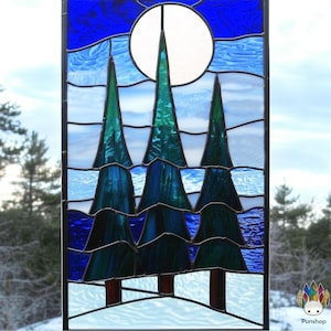 Puede incluir: Una ventana de vitrales que representa una escena de invierno con tres árboles de hoja perenne y una luna llena en un cielo azul.