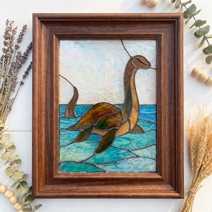 May include: A stained glass panel in a dark brown wooden frame depicting a long-necked creature swimming in blue water. The creature is rendered in shades of brown and green glass, while the water features textured blue and teal glass pieces.