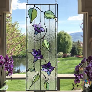 Peut inclure: Panneau rectangulaire en vitrail avec un motif floral. Il présente des fleurs violettes et des feuilles vertes sur une vigne, sur un fond de verre texturé. Le panneau est encadré de métal noir et suspendu à une chaîne.