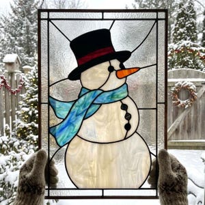 Pode incluir: Um painel retangular de vitral com um boneco de neve usando cartola preta, faixa vermelha, nariz de cenoura laranja e cachecol com tons de azul e verde. O boneco tem três botões pretos sobre um fundo de vidro transparente.