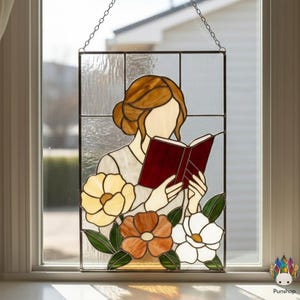 May include: Stained glass panel featuring a woman reading a book, with a floral design. The woman has brown hair and is wearing a light-colored top. The flowers are yellow, orange, and white, with green leaves. The panel is hanging by a chain.