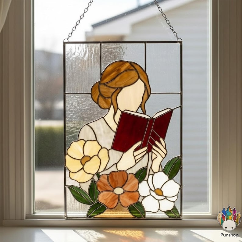 May include: Stained glass panel featuring a woman reading a book, with a floral design. The woman has brown hair and is wearing a light-colored top. The flowers are yellow, orange, and white, with green leaves. The panel is hanging by a chain.