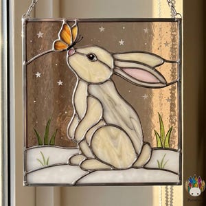 Op de afbeelding: Gebrandschilderd glaspaneel met een witte konijn met roze oren, kijkend naar een oranje vlinder. De achtergrond is een bruine gradiënt met witte sterren. Groen gras en witte sneeuw onderaan.