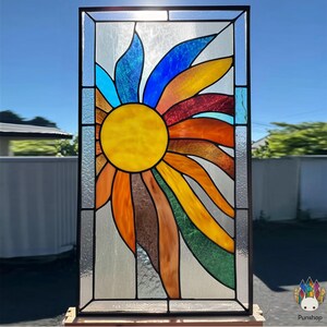 May include: A colourful stained glass panel depicting a sunflower. The central sun is yellow, surrounded by petals in orange, red, blue, and green. The panel is framed in black and set against a clear blue sky.
