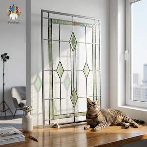 Puede incluir: Una ventana decorativa de vidrieras con un diseño geométrico con formas de diamantes verdes. La ventana está enmarcada en un metal plateado y está al lado de una ventana. Un gato atigrado está acostado sobre una superficie de madera.