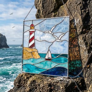 May include: A square stained glass panel featuring a red and white striped lighthouse on a tan cliff, a small sailboat on blue and teal waves, and two white birds in a light blue sky, all held together by a metal frame and hanging chains.