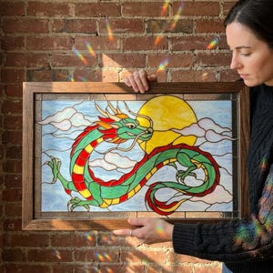 Puede incluir: Un panel rectangular de vidrieras en un marco de madera que presenta un dragón verde con detalles en rojo y amarillo. El dragón está posicionado sobre un fondo de un gran sol amarillo y nubes azul claro. El vidrio tiene un acabado texturizado.