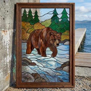 May include: A rectangular stained glass panel in a dark wood frame depicting a brown bear walking through a shallow river. The scene includes green pine trees, textured amber foliage, and blue water with grey rocks, all crafted from various glass pieces.