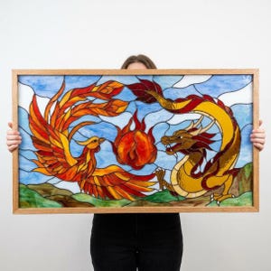 Puede incluir: Un panel de vidriera rectangular en un marco de madera que representa un fénix con alas naranjas y rojas frente a un dragón amarillo. Entre ellos hay una esfera brillante roja y naranja. El fondo muestra un cielo azul claro y colinas verdes.