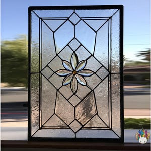 May include: Stained glass window panel in a black frame. The design includes a central flower with clear and amber petals, set within geometric shapes. The glass has a textured, rippled look, and the background shows an outdoor view.