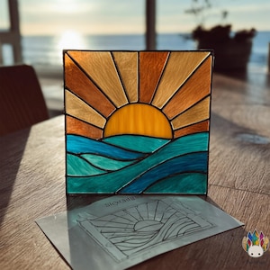 May include: A square stained glass panel depicting a sunset over ocean waves. The sun is a vibrant yellow, with orange and tan rays. The waves are shades of teal and blue. A paper with a line drawing of the design is in front of the panel.