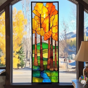 Pode incluir: Um painel de janela de vitral com uma paisagem de outono estilizada. O design inclui árvores com folhagem amarela e laranja, troncos marrons, grama verde e um riacho azul. O painel é emoldurado em preto e colocado em frente a uma janela.