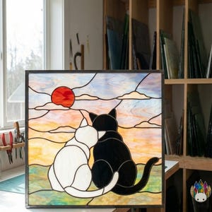 May include: Stained glass panel featuring two cats, one white and one black, gazing at a sunset with a red sun and clouds. The artwork is framed in a dark border and set against a background of a workshop.