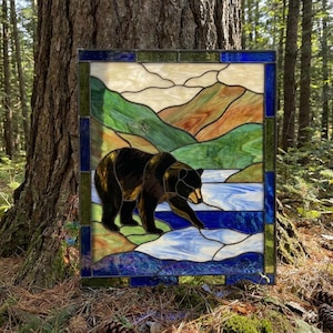 May include: A rectangular stained glass panel featuring a black bear walking along a blue riverbank. The background depicts rolling green and brown hills under a cream-coloured sky. The entire scene is framed by a dark blue and green glass border.