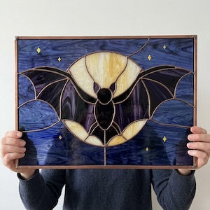 May include: A rectangular stained glass panel featuring a black bat silhouette centered against a large, pale yellow circular moon. The background is composed of deep blue swirled glass with small, scattered yellow diamond-shaped glass stars.