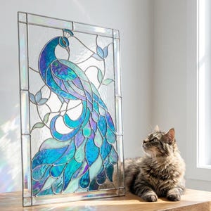 以下が含まれることがあります： 青、ターコイズ、紫の色合いの孔雀を描いたステンドグラスパネル。 孔雀は緑の葉に囲まれ、長方形のフレームに入っています。 パネルの隣には、縞模様の猫が横たわっています。