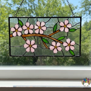 May include: Stained glass panel depicting a branch with pink cherry blossoms and green leaves. The flowers have yellow centres and are set against a clear background. The panel is framed in black and hangs from a chain.