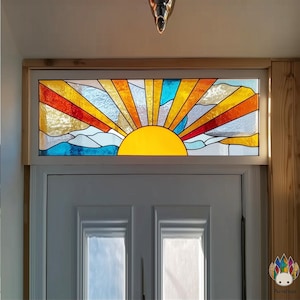 May include: A stained glass window featuring a sunrise design. The sun is a bright yellow, with rays in orange, red, and yellow. The background includes blue and white clouds. The window is set in a white frame above a white door.