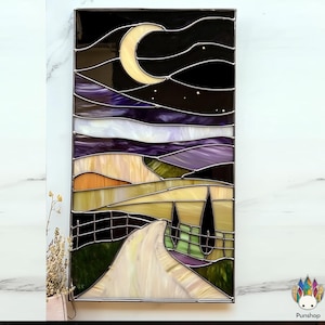 Può includere: Una finestra in vetro colorato che rappresenta una scena notturna con una luna crescente, stelle e una strada tortuosa che conduce a un campo con due alberi. Il cielo è un gradiente di viola, blu e bianco, e il campo è un mix di verde, giallo e arancione.