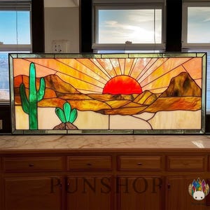 May include: Stained glass panel depicting a desert landscape with a setting sun. The artwork features a vibrant red sun, mountains, and cacti in shades of orange, yellow, and green. The panel is framed with a dark border.