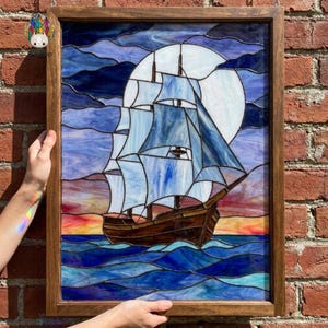 May include: A rectangular stained glass panel in a wooden frame depicting a sailing ship on blue waves. The ship features white and blue sails against a large white moon. The background shows a sunset sky with orange, pink, and dark blue glass.