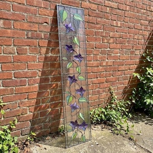 Op de afbeelding: Een hoog, rechthoekig glas-in-loodpaneel met een motief van een klimplant met paarse bloemen en lichtgroene bladeren. Het ontwerp is geplaatst tegen een heldere, getextureerde glazen achtergrond, omlijst door een dunne metalen rand verticaal.