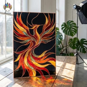 May include: A stained glass panel featuring a phoenix design. The bird is depicted with vibrant colors including red, orange, yellow, and green against a black background. The artwork is framed with a copper border and is displayed on a wooden surface.