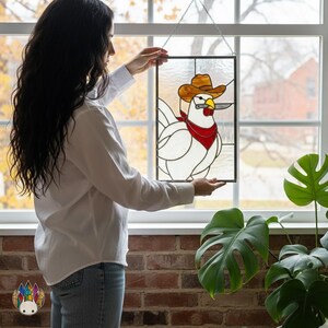 Könnte beinhalten: Buntglasfenster mit einem weißen Huhn, das einen Cowboyhut und ein rotes Halstuch trägt und ein Messer hält. Das Kunstwerk ist schwarz gerahmt und hängt an einer silbernen Kette. Der Hintergrund zeigt ein Fenster mit Herbstlaub.