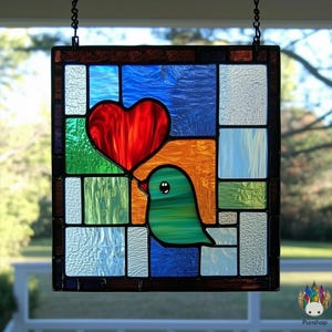 Può includere: Un pannello di vetro colorato con un uccello verde che tiene un cuore rosso nel becco. Il pannello è composto da quadrati colorati in tonalità di blu, verde, arancione e rosso.