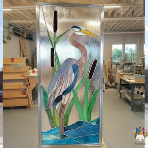 May include: Stained glass panel depicting a great blue heron in a wetland scene with cattails. The heron has blue, grey, and brown plumage, a yellow beak, and a white head stripe. The panel is framed in silver metal.